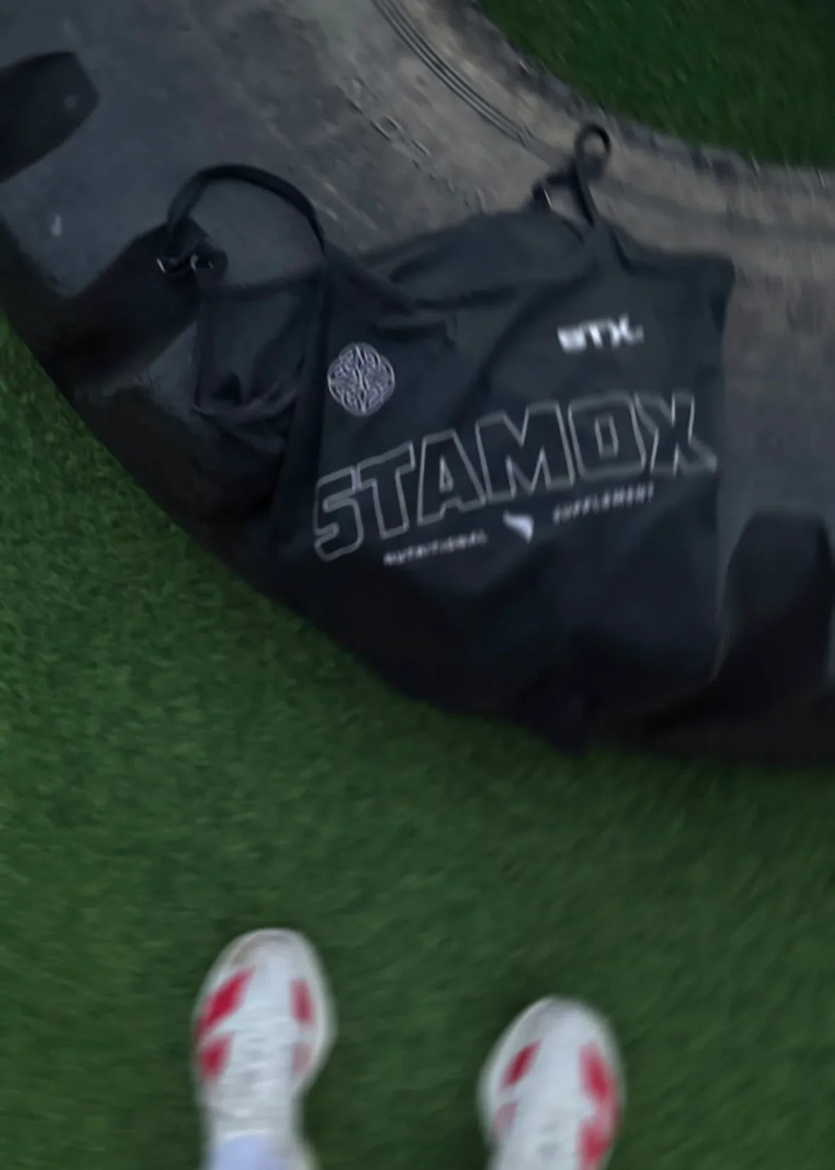 Person wearing white sneakers with red accents on a grassy field, standing next to a black bag with 'stamox' branding.