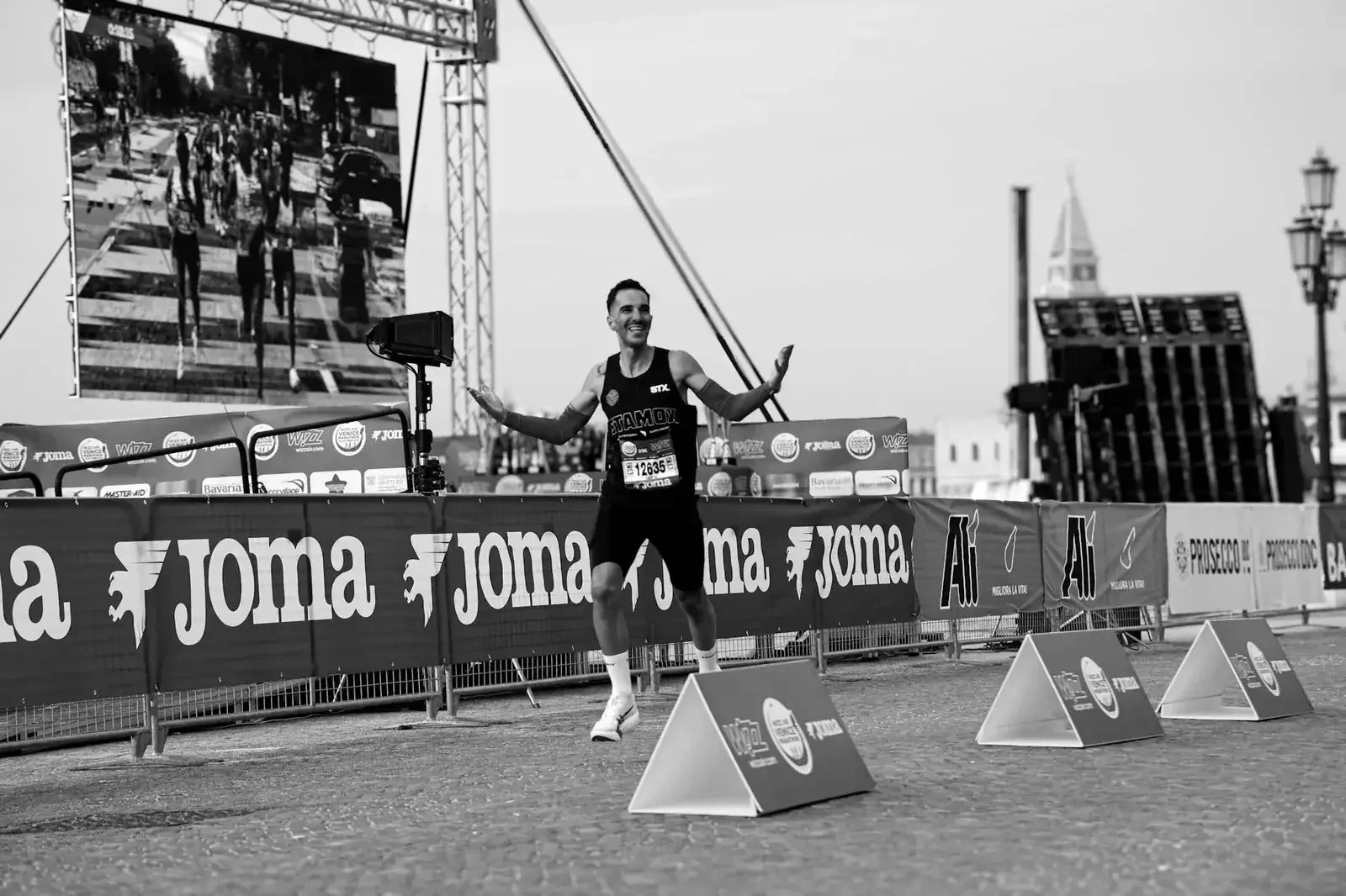 Runner celebrating finish line with Stamox on race bib, Joma banners, endurance event