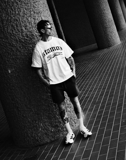 Man wearing stamox beetroot powder t-shirt, shorts, and sneakers, standing by pillar