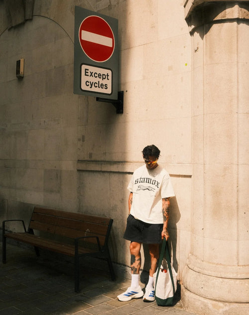 Man wearing stamox beetroot powder t-shirt, standing by a wall with a tote bag and street signs.