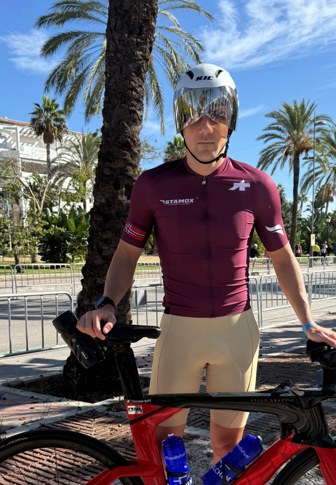 Man with a bicycle in an outdoor setting with palm trees and buildings in the background