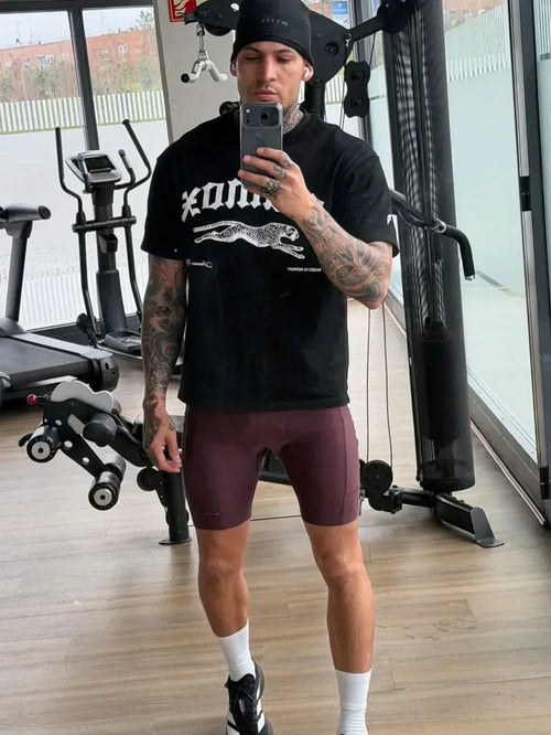 Athletic man taking mirror selfie at gym wearing black t-shirt, gym shorts, promoting stamox beetroot powder