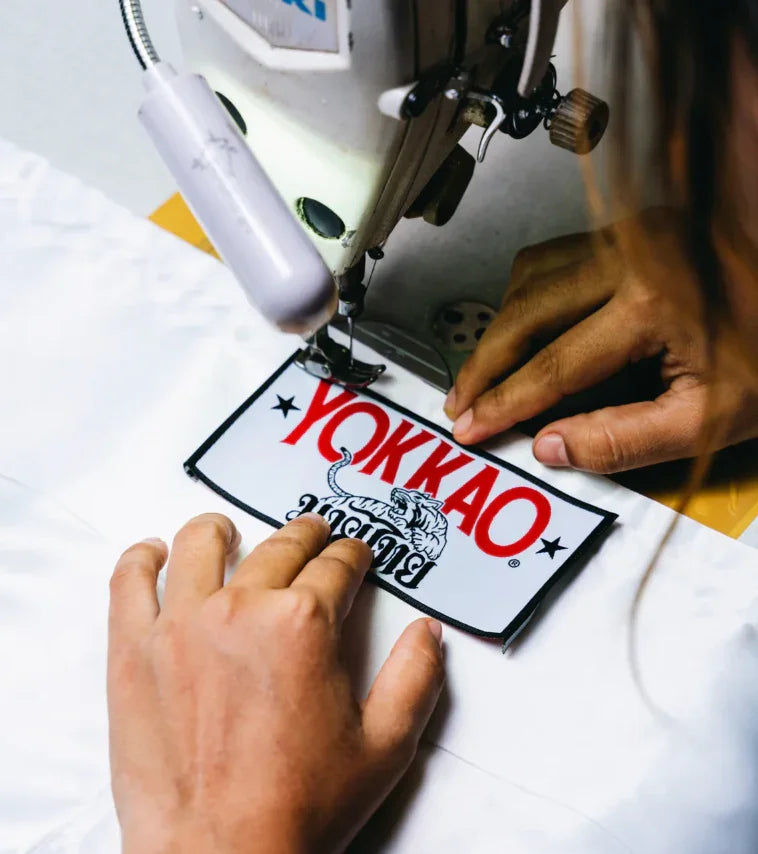 Hands sewing YOKKAO logo patch on white fabric with industrial machine