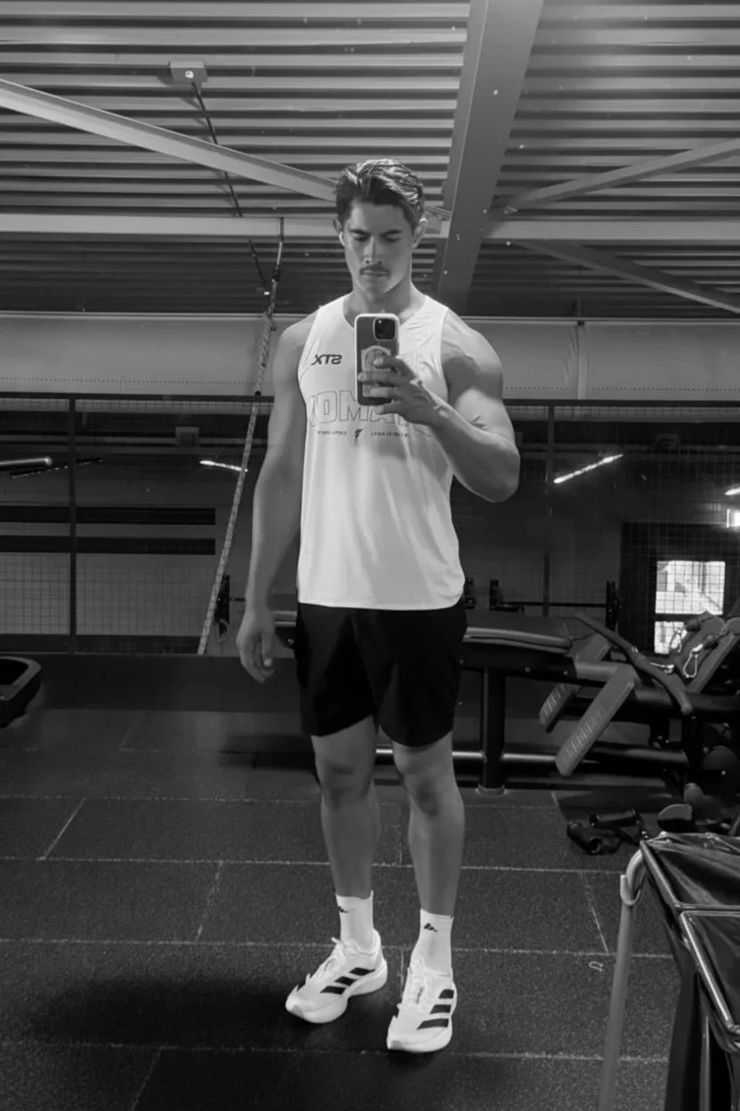 A man takes a mirror selfie in a gym wearing a white athletic tank top with 'A10' printed on the chest, black shorts, white socks, and white running shoes. stamox STX white singlet running.