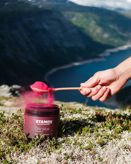 Hand scooping vibrant beetroot powder from Stamox Oxy-plus jar atop mountain, boosting endurance & recovery naturally.