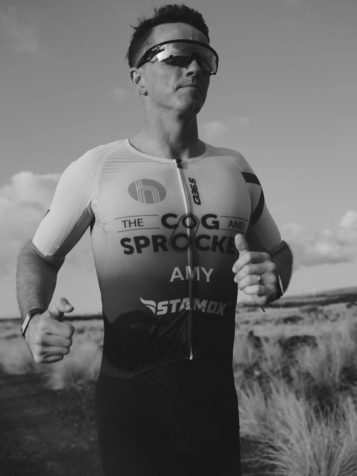 A man wearing a cycling jersey and sunglasses outdoors in a black and white photograph. The jersey has visible sponsor logos including Cog and Sprock, Amy, and StamoX.