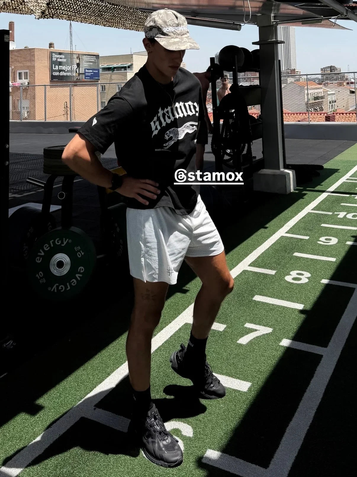A man wearing a black Stamox t-shirt, white athletic shorts, and black sneakers stands on a green turf track in a gym setting. The image features a visible '@stamox' watermark overlay.