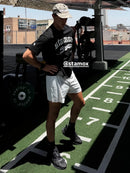 A man wearing a black Stamox t-shirt, white athletic shorts, and black sneakers stands on a green turf track in a gym setting. The image features a visible '@stamox' watermark overlay.
