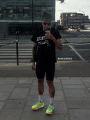 A man takes a mirror selfie in an urban setting, wearing a black graphic t-shirt, black athletic shorts, white socks, and neon yellow running shoes.