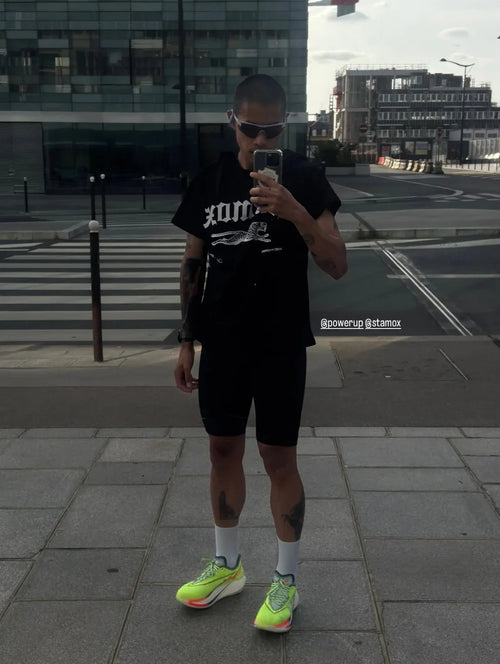 A man takes a mirror selfie in an urban setting, wearing a black graphic t-shirt, black athletic shorts, white socks, and neon yellow running shoes.