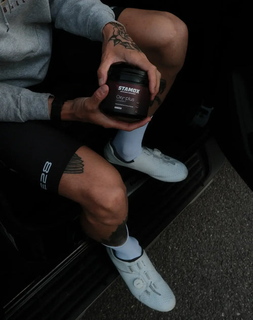 Cyclist holding Stamox beetroot powder Oxy-plus, wearing cycling gear and white shoes.
