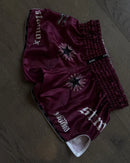 Maroon shorts with floral embroidery and branding on a wooden surface. STAMOX YOKKAO SHORTS