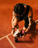 A tattooed man crouches on an orange running track, preparing a drink by opening a black shaker bottle and a red supplement tub. He wears a black tank top and shorts, with white socks and a white sneaker.