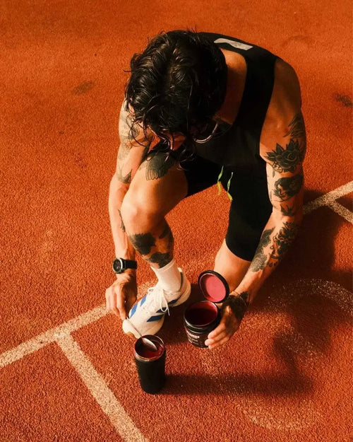 A tattooed man crouches on an orange running track, preparing a drink by opening a black shaker bottle and a red supplement tub. He wears a black tank top and shorts, with white socks and a white sneaker.